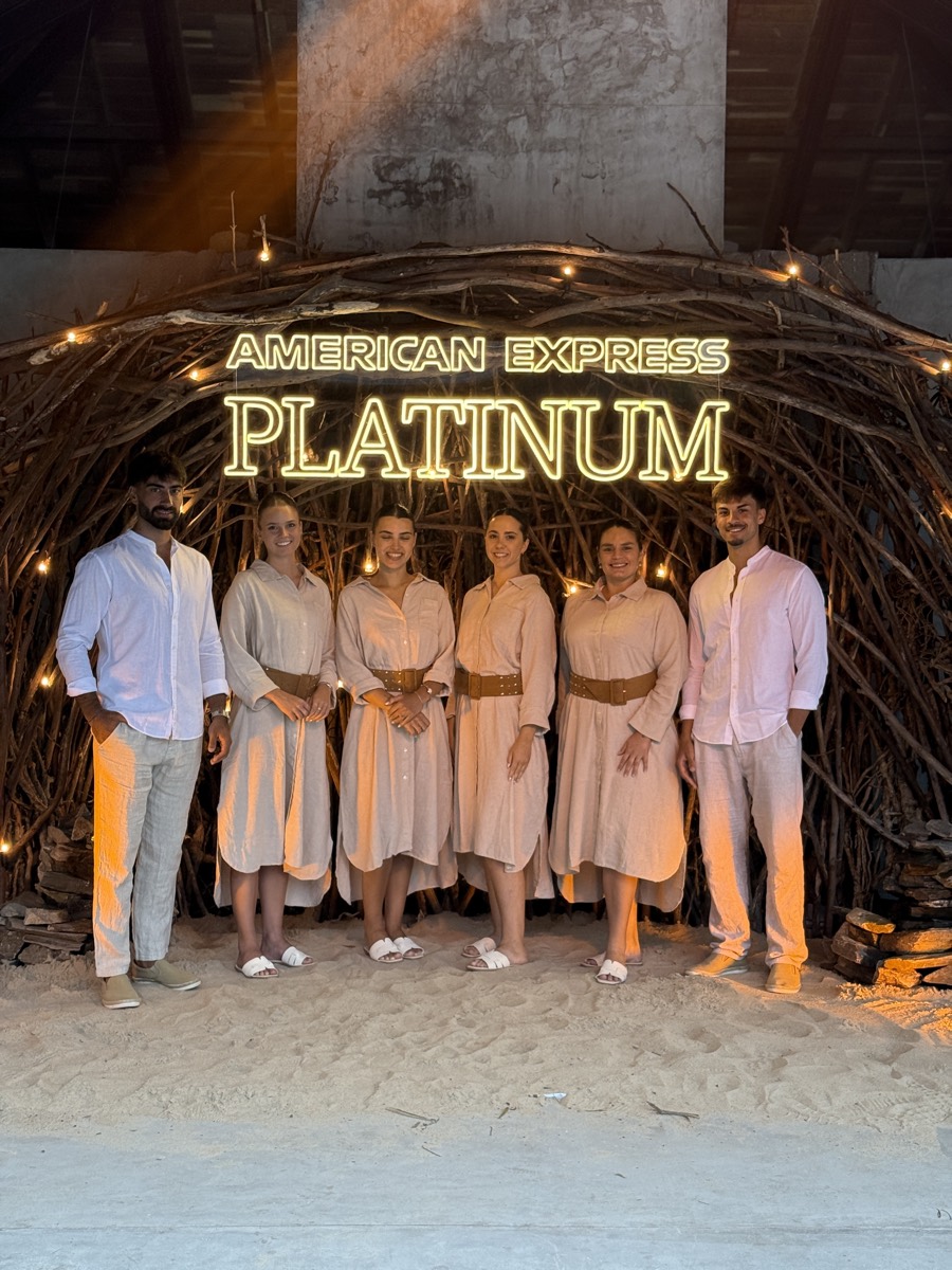 Equipo de anfitriones en tonos durazno frente a un arco de ramas con el cartel American Express Platinum.