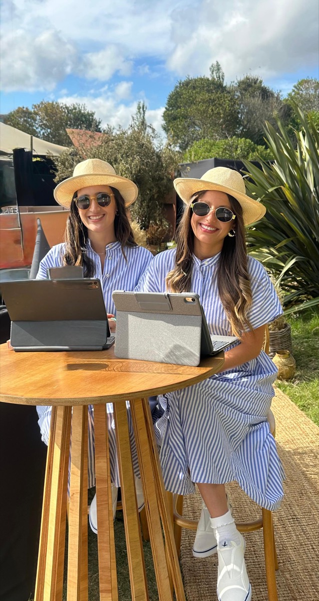 Dos anfitrionas con sombrero en la mesa de registro de un evento al aire libre.